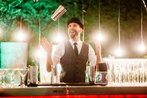 Vanilla Bean Catering flair bar tender throwing a mixing glass while standing behind a bar illuminated with festoon lighting
