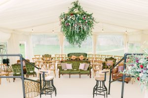 Marquee Furniture. A large floral decoration hanging over a sofa, chairs and other furniture laid out inside a white lined marquee for a event.