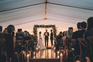 Winter weddings. A couple standing at the alter in a marquee, viewed fron the rear of the aisle with guests seated either side of the aisle. Wedding marquee hire